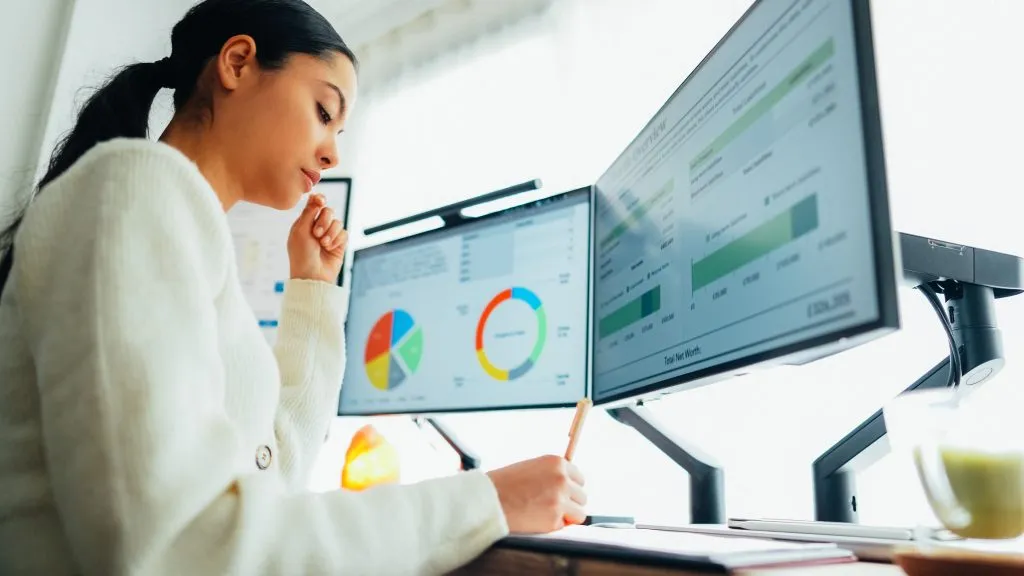 A woman in a white sweater focuses intently on two computer screens displaying marketing agency performance